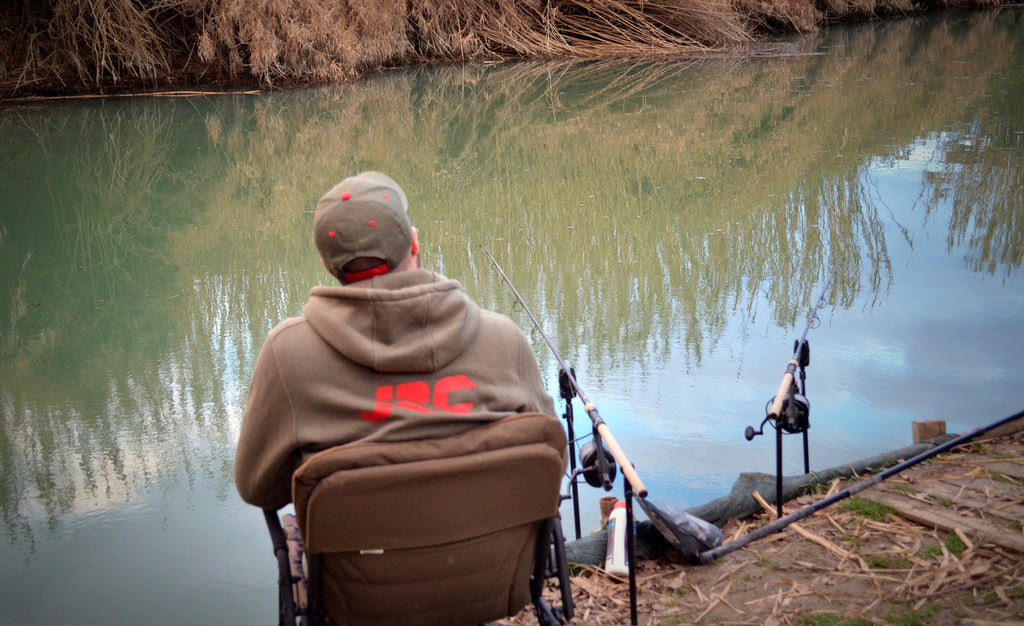 A person sitting in a folding fishing chair, viewed from behind, wearing a brown hoodie with a red 'JRC' logo and a matching cap, fishing by a calm river with reeds in the background.
