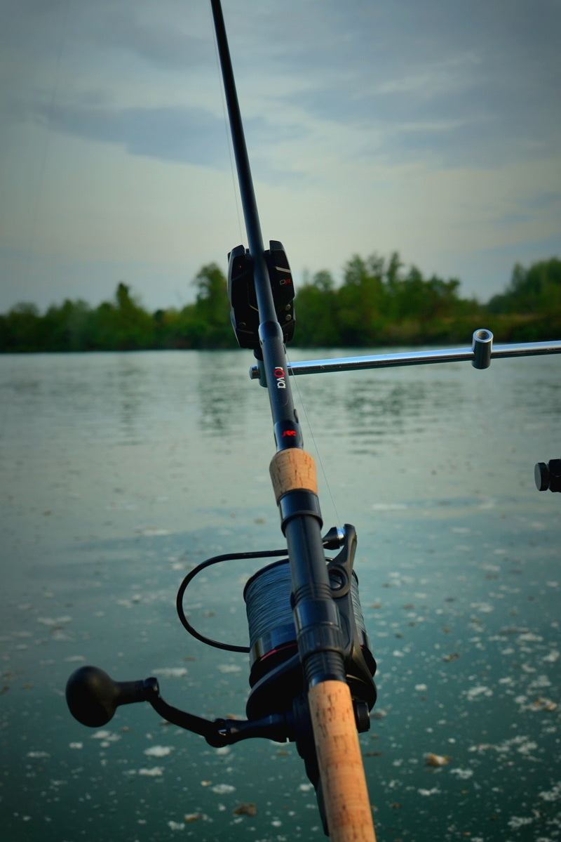 A close-up of a fishing rod and spinning reel mounted on a stand, with a calm lake and tree-lined shore in the background.