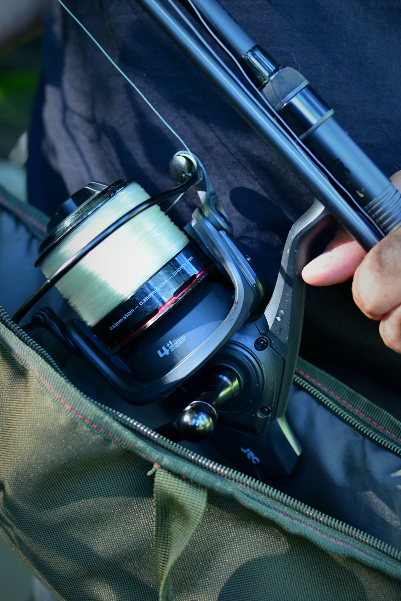 A close-up of a black spinning fishing reel mounted on a rod, partially tucked inside an olive green bag, with a hand holding the rod.